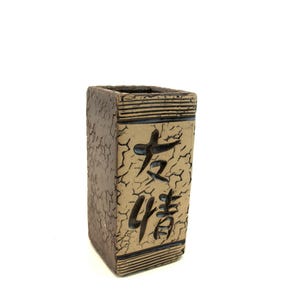 May include: A rectangular, brown ceramic vase with a textured surface and black accents. The vase features Japanese characters on the front. The top and bottom edges have horizontal black lines.