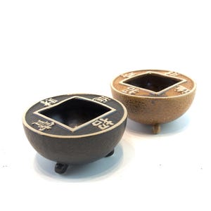 May include: Two small, round, decorative bowls with a coin-like design. One is black with a silver border, the other is bronze. Each has a square opening in the center and rests on three small feet.
