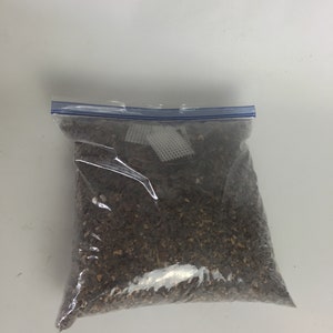 May include: A clear plastic bag filled with brown and tan organic material, possibly mulch or potting mix. The bag is sealed with a blue zipper closure and sits against a white background.