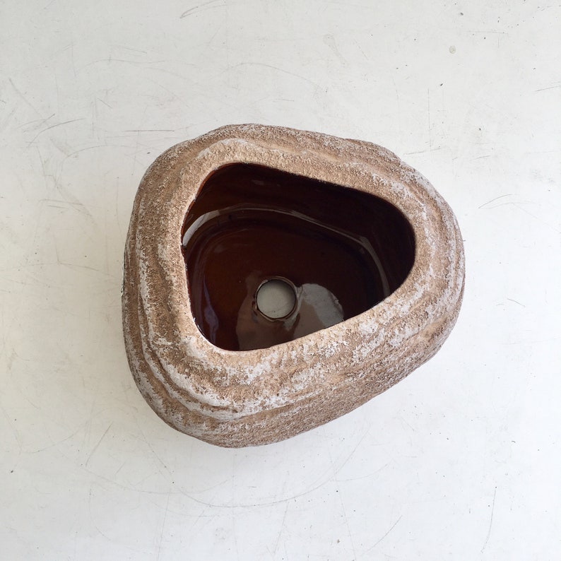 May include: A brown ceramic sink basin with a textured, stone-like exterior. The interior is a glossy dark brown, and the basin has a unique, organic shape. The sink has a single drain hole in the center.