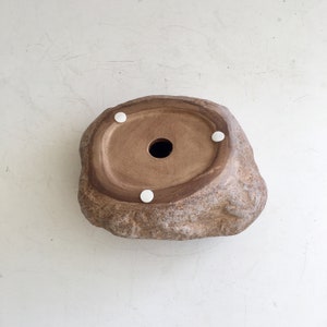 May include: A brown ceramic planter shaped like a rock with a drainage hole in the center. The planter has four white rubber feet on the bottom.