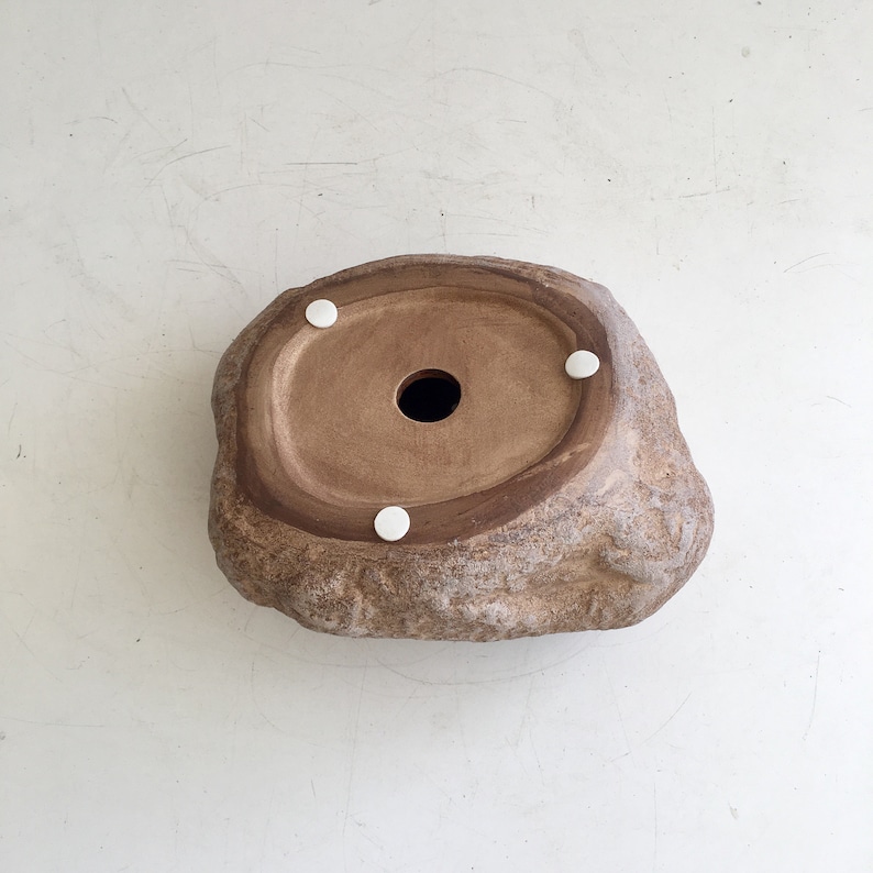 May include: A brown ceramic planter shaped like a rock with a drainage hole in the center. The planter has four white rubber feet on the bottom.