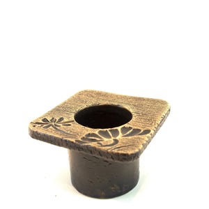 May include: A brown, square-shaped bottle stopper with a floral design carved into the top. The stopper has a cylindrical base and a textured surface, suitable for a decorative touch.