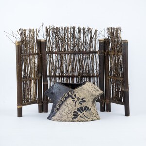 May include: A decorative ceramic vase with a black and white design featuring stylized flowers and swirls. The vase is sitting in front of a bamboo fence.