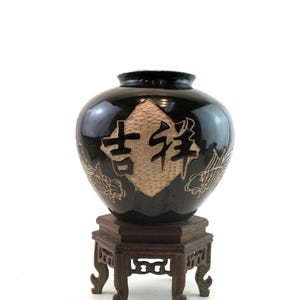 May include: A black ceramic vase with gold Chinese characters and dragon motifs, displayed on a dark wooden stand. The vase has a glossy finish and a rounded shape, showcasing traditional Asian design.