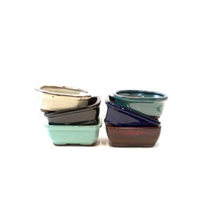 May include: A collection of small, rectangular ceramic bonsai pots in various colors. The pots are stacked in two groups, showcasing shades of cream, black, teal, navy, and burgundy. The pots have a simple, minimalist design.