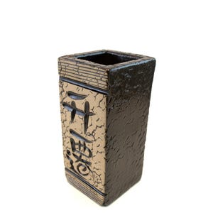May include: A rectangular ceramic vase with a textured, dark brown exterior and a lighter tan front panel. The front panel features black Japanese characters. The top rim has a dark brown glaze, and the vase has a square opening.