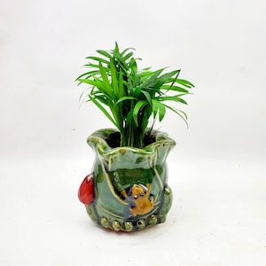 May include: A green ceramic planter shaped like a frog with a red accent and a small green plant growing inside.