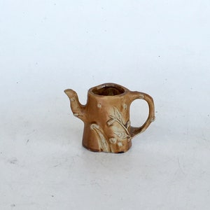 May include: A small brown ceramic teapot with a tree trunk design and a floral pattern. The teapot has a spout and a handle.