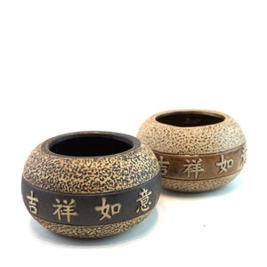 May include: Two round, ceramic bowls with a textured, speckled surface. Each bowl has a dark band with Chinese characters. The bowls are brown and beige, and the interior is dark. The bowls are likely for decorative or functional use.