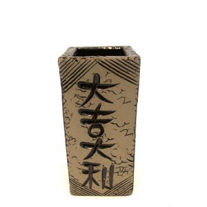 May include: A rectangular, beige ceramic vase with black Japanese characters. The vase has a textured surface and a dark interior. The characters are vertically aligned and appear to be hand-painted. The vase is a decorative item.