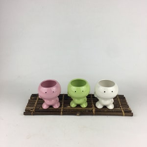 May include: Three small ceramic planters shaped like cartoon characters with smiling faces. The planters are pink, green, and white and are sitting on a brown bamboo mat.