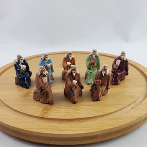 May include: Eight small ceramic figurines of elderly men with white beards, wearing colorful robes and holding various objects, are arranged on a round wooden tray.