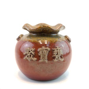 May include: A small, round, ceramic jar with a ruffled rim and two small handles. The jar is primarily reddish-brown with a band of brown and gold near the top. The jar has Chinese characters embossed on the front.
