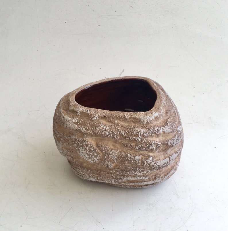 May include: A decorative, textured, stone-like planter with a wide, oval opening. The exterior is a light brown with white accents, and the interior is a dark brown. The planter has a unique, organic shape, suitable for indoor or outdoor use.