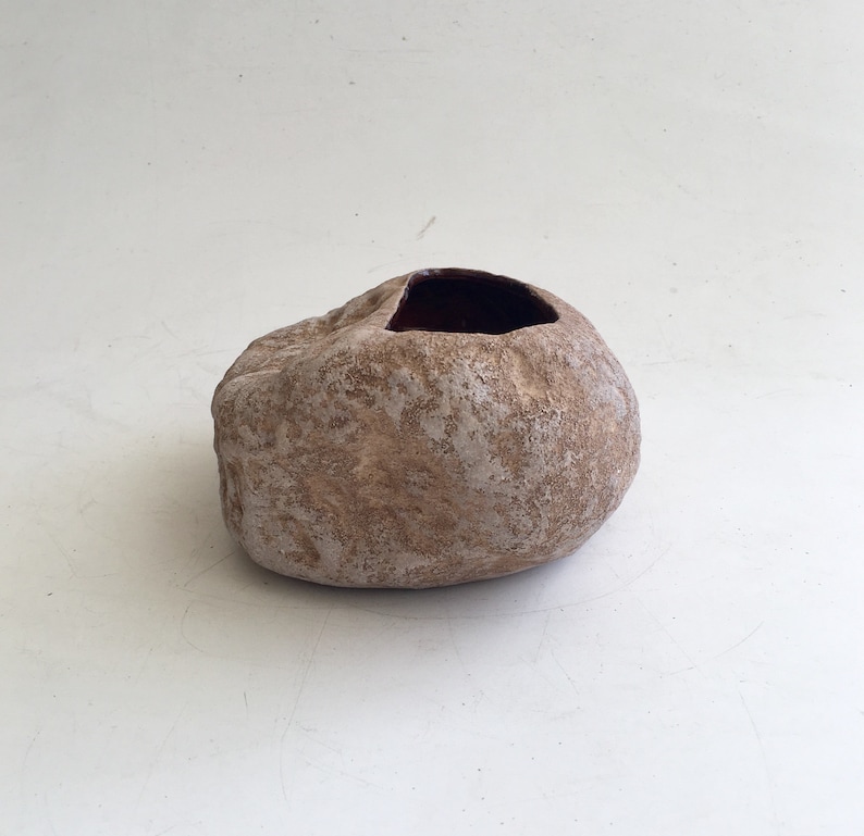 May include: A small, round, brown ceramic planter with a rough, textured surface. The planter has a wide, open top and a slightly uneven rim.