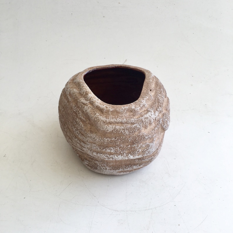 May include: A small, textured, brown and white ceramic pot with a dark interior. The pot has a unique, organic shape with a wide opening. The exterior features a layered, ridged design, creating a rustic appearance. Ideal for small plants or decorative use.