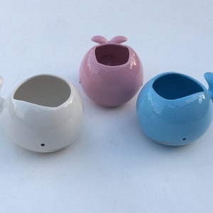 Op de afbeelding: Drie keramische walvisvormige plantenbakken in wit, roze en blauw. Elke plantenbak heeft een ronde opening voor planten, een staartvin en een kleine zwarte stip voor een oog. De plantenbakken zijn geschikt voor kleine vetplanten.