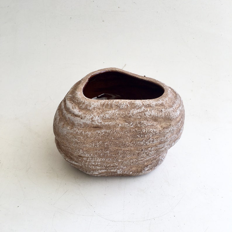 May include: A small, brown ceramic planter with a textured, stone-like appearance. The planter has a wide, irregular opening and a lighter, almost white, color highlights the textured surface. The interior is a darker brown.