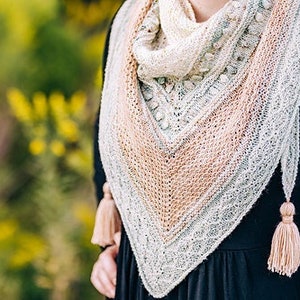May include: A knitted shawl in a triangular shape, featuring a gradient of colors from cream to peach to gray. The shawl has a delicate lace pattern and is adorned with tassels at the ends. The person is wearing a black dress.