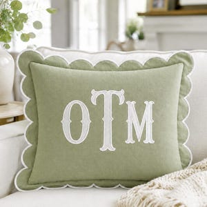 Op de afbeelding: Een saliegroen rechthoekig sierkussen met een geschulpte rand en witte afwerking. In het midden van het kussen staan de geborduurde witte monogramletters OTM in een schreeflettertype. Het kussen heeft een effen kleur en een decoratieve rand.