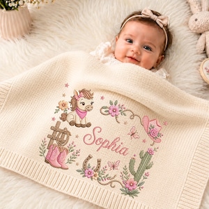 Könnte beinhalten: Eine cremefarbene Strickdecke für Babys mit einer Stickerei, die ein Pony, rosa Cowboystiefel, ein Lasso, ein Hufeisen, einen Kaktus und einen rosa Cowboyhut zeigt. Der Name Sophia ist in rosa Schreibschrift in der Mitte der Decke eingestickt.