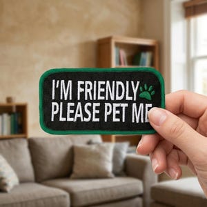 May include: A rectangular black patch with a green border and white text that reads "I'M FRIENDLY PLEASE PET ME." A green paw print is on the right side of the text. The patch is held by a hand.
