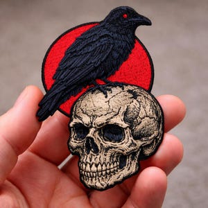 May include: Embroidered patch featuring a black raven with red eyes perched atop a skull. The raven is set against a red circle, with the skull in a tan color. The patch is held in a hand.