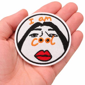 May include: A round, white embroidered patch with a cartoon face design. The patch features black eyebrows and hair, orange text that reads "I am cool", and a red lip design.