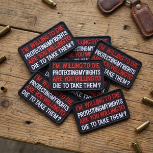 May include: A collection of black rectangular patches with white and red text. The text on each patch reads "I'M WILLING TO DIE PROTECTING MY RIGHTS ARE YOU WILLING TO DIE TO TAKE THEM?" The patches are on a wooden surface.