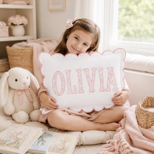 Puede incluir: Un cojín decorativo rectangular blanco con borde festoneado rosa, que presenta el nombre OLIVIA bordado en letras mayúsculas grandes de color rosa con estampado floral. El cojín es sostenido por un niño, con un peluche de conejo blanco cerca.