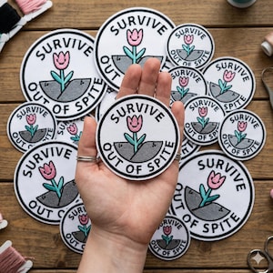 May include: A collection of white, circular patches with the text "SURVIVE OUT OF SPITE" in black. Each patch features a pink tulip with green stem and leaves. The patches are on a wooden surface.