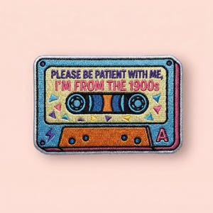 May include: Embroidered patch in the shape of a cassette tape. The patch is blue, orange, and yellow with the text "PLEASE BE PATIENT WITH ME, I'M FROM THE 1900s". The cassette tape design includes details like the tape reels and the "A" label.