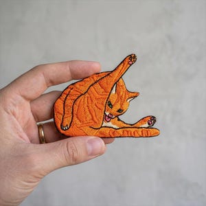 May include: Embroidered patch featuring an orange cat grooming itself. The cat is depicted in a playful pose, with its leg raised and tongue sticking out. The patch has detailed stitching and a black outline.