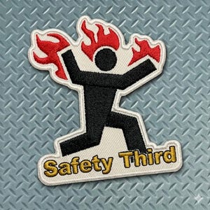 May include: Embroidered patch featuring a black stick figure with arms raised and flames above the head. The patch has a white border and the words "Safety Third" in gold lettering.