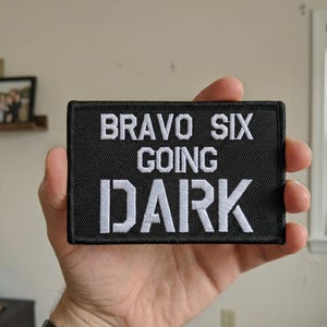 May include: Black rectangular patch with white embroidered text that reads "BRAVO SIX GOING DARK". The patch has a black border and is held in a hand.