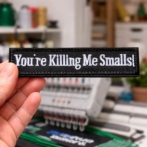 May include: Black rectangular embroidered patch with the phrase "You're Killing Me Smalls!" in white lettering. The patch is held in a person's hand, with an embroidery machine in the background.