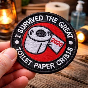 May include: A round, black patch with white text that reads "I SURVIVED THE GREAT TOILET PAPER CRISIS". The center features a red circle with a white roll of toilet paper and the year "2020".