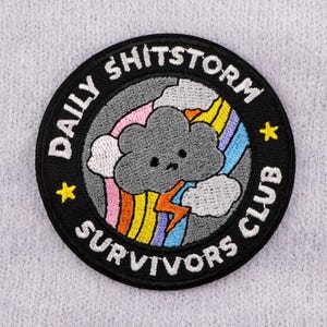 May include: Embroidered patch with the text "DAILY SHITSTORM SURVIVORS CLUB" in white on a black circular background. The patch features a gray cloud with a sad face, a rainbow, and a yellow lightning bolt.