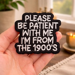 May include: Black embroidered patch with white text that reads "PLEASE BE PATIENT WITH ME I'M FROM THE 1900'S". The patch has a black border and is held in a hand.