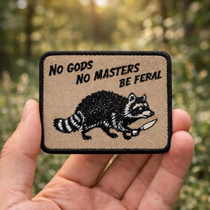 May include: A rectangular patch with a tan background and black border. The patch features a black and white raccoon holding a knife, with the text "No Gods No Masters Be Feral" in black.