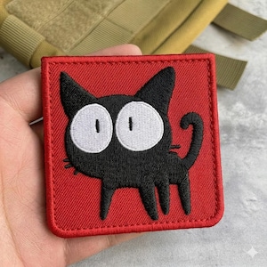 May include: A square, red patch featuring a black cartoon cat with large white eyes. The cat has a curved tail and is embroidered with black thread. The patch is likely for clothing or accessories.
