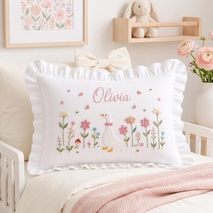 Puede incluir: Una almohada rectangular blanca con borde de volantes, que presenta detalles bordados de una oca blanca, flores coloridas y una pequeña seta. El nombre Olivia está bordado en cursiva rosa en el centro superior, junto a pequeñas mariposas.