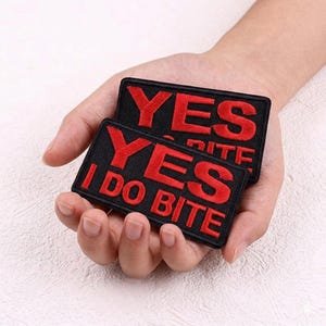 May include: Two black rectangular embroidered patches with red text. The top patch reads "YES" and the bottom patch reads "YES I DO BITE". The patches are held in a hand.