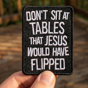 May include: Black embroidered patch with white text that reads "DON'T SIT AT TABLES THAT JESUS WOULD HAVE FLIPPED." The patch is rectangular with rounded corners and is held by a hand.