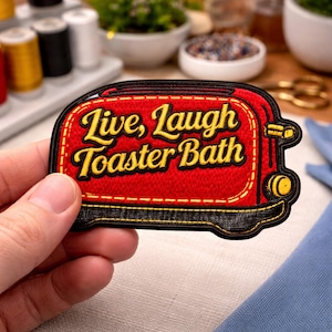 May include: A red and black embroidered patch in the shape of a toaster. The patch features the words "Live, Laugh, Toaster Bath" in yellow script. The patch has a black border and yellow accents.