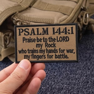 May include: A rectangular patch with the text "PSALM 144:1 Praise be to the LORD my Rock who trains my hands for war, my fingers for battle." The patch is tan with black text and a black border. The background is a tan tactical bag.