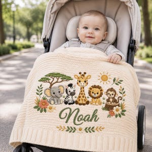 May include: A cream-colored knitted baby blanket featuring an embroidered safari scene with an elephant, zebra, giraffe, lion, and monkey. The name Noah is embroidered in green script below the animals, surrounded by trees, flowers, and a sun.