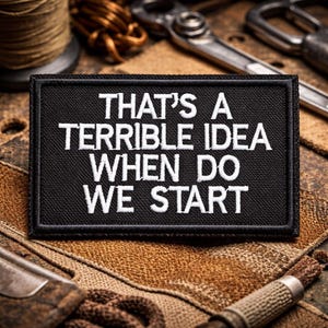 May include: Black rectangular patch with white embroidered text: "THAT'S A TERRIBLE IDEA WHEN DO WE START." The patch is surrounded by a black border. The background includes tools and spools of thread.