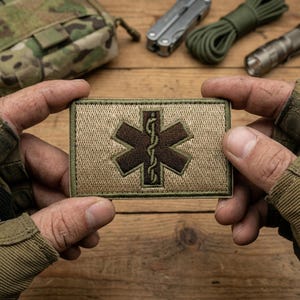 May include: A rectangular patch with a brown medical symbol on a tan background, framed by a green border. The patch is held between two hands, with other survival gear in the background.